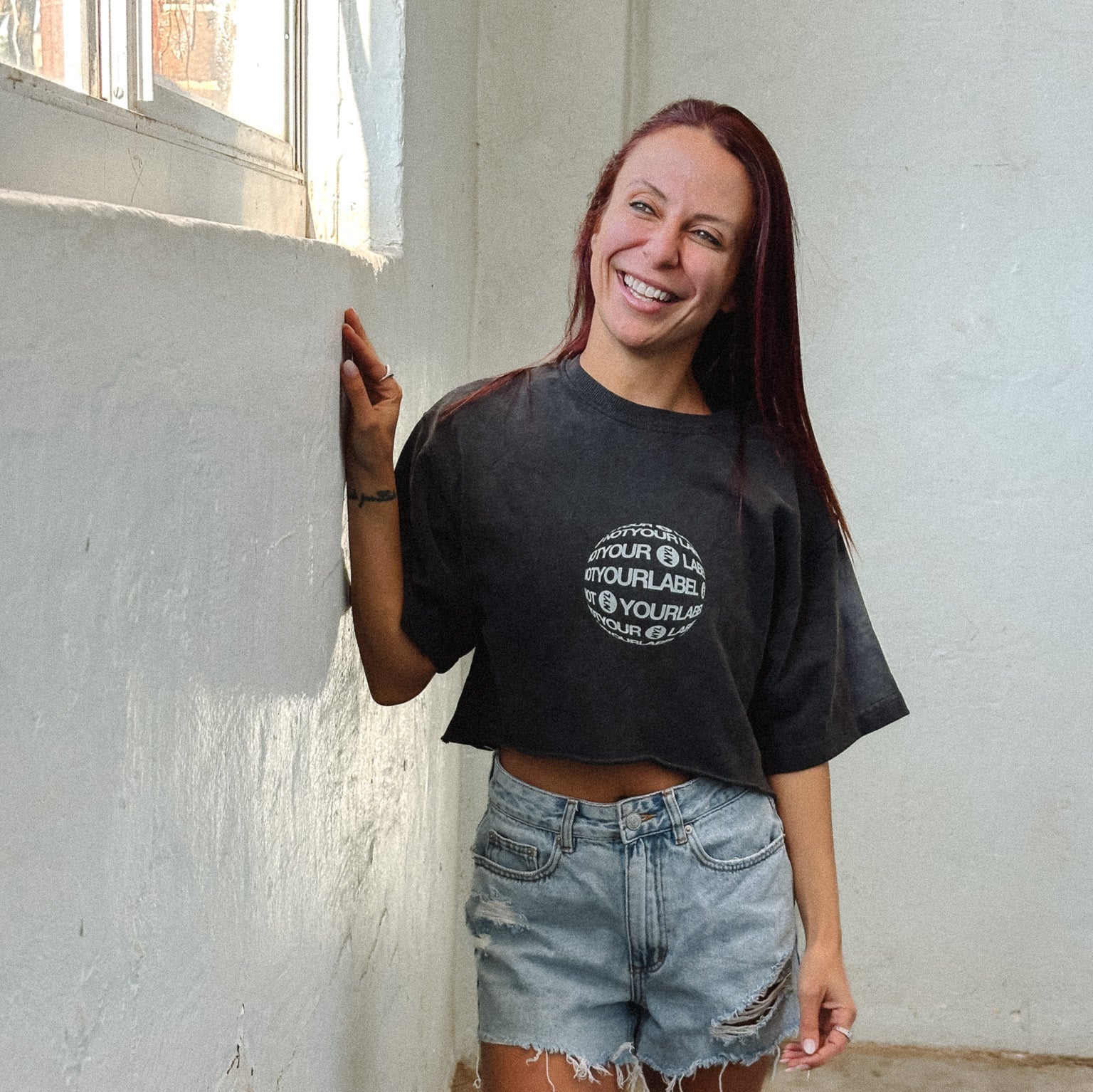 Woman wearing a grey oversized Not Your Label t-shirt and denim shorts standing against a white wall.